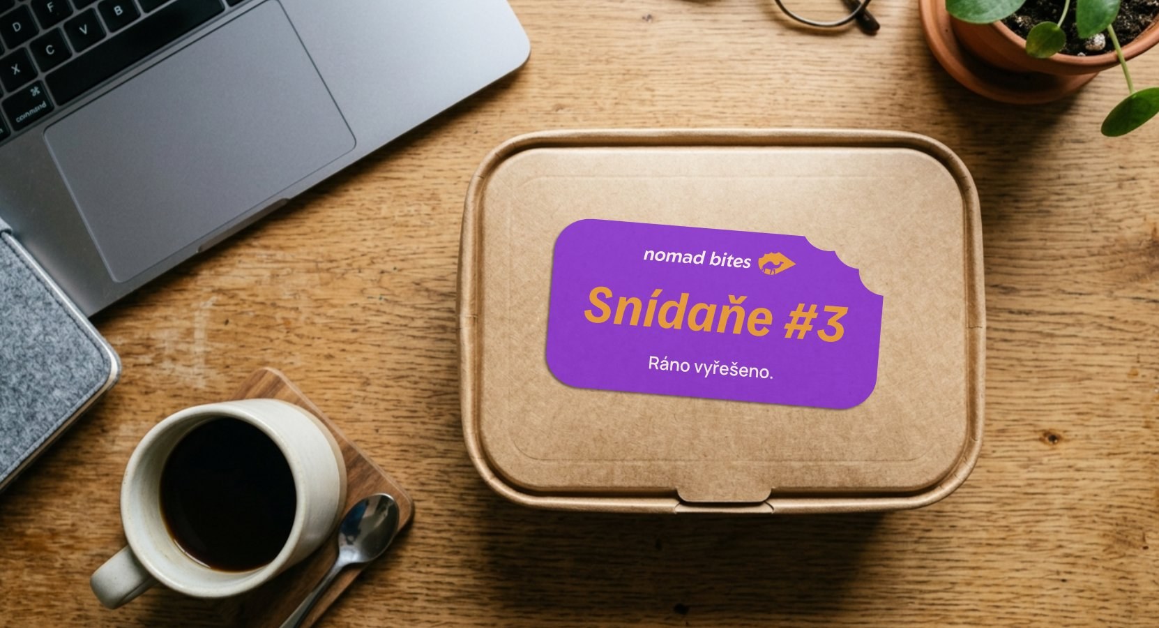 Krabička Nomad Bites se samolepkou na pracovním stole vedle notebooku a kávy